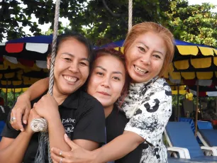 DSCN1029.JPG: Three friends sharing a warm group hug and bright smiles outdoors, with colorful umbrellas and lounge chairs in the background.