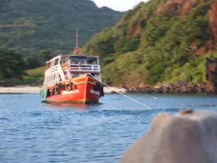 DSCN1066.JPG: A red wooden boat anchored in calm blue water near a lush green shoreline, with hills rising in the background and a sandy beach visible to the left.
