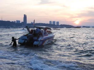 DSCN1073.JPG: A speedboat pulls up to a sandy shore at sunset as people climb aboard, the city skyline silhouetted in the distance.