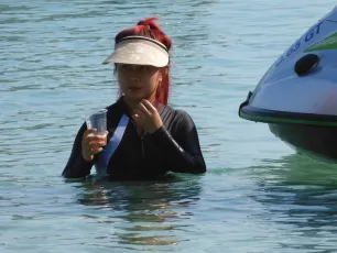 DSCN0944.JPG: A woman in a visor and wetsuit stands waist-deep in calm blue water, holding a clear plastic cup near a jet ski.