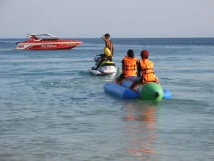DSCN1009.JPG: Two people in orange life jackets ride a blue inflatable banana boat being towed by a jet ski, with a red speedboat visible in the distance.