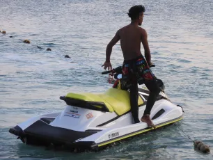 DSCN1054.JPG: A young man in patterned swim trunks stands on the back of a yellow-and-white Yamaha jet ski as it floats in calm, shallow water.