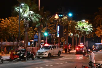 DSC_3274: Nighttime street scene with palm trees, parked vehicles and scooters, and pedestrians passing beneath traffic lights and street lamps.