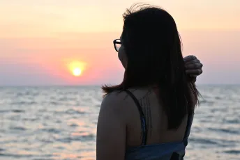 DSC_3513: A woman in glasses watches the sun dip below the horizon over a calm sea, silhouetted against a pastel sunset.