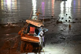 DSC_3654: A small boat listing on a muddy shoreline at low tide, its hull illuminated by nearby lights reflecting on the water.