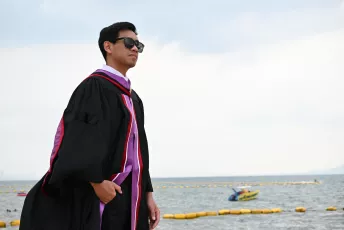 DSC_3816: A graduate in gown and sunglasses stands confidently by the water, looking toward the horizon on a bright day.