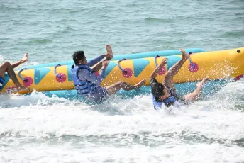 DSC_4881: Two riders tumble into the surf as a colorful inflatable banana boat capsizes during a lively ocean ride.