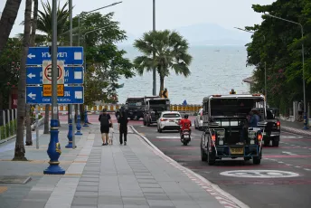 DSC_4764: A seaside road with palm trees and directional signs, vehicles and motorbikes heading toward the waterfront while pedestrians walk along the paved sidewalk.