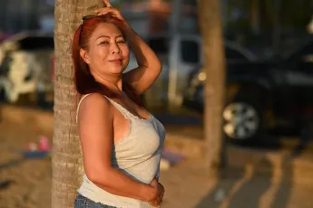 DSC_1717: A woman in a gray tank top leans against a palm tree on a sunlit beach, smiling as she enjoys the warm afternoon.