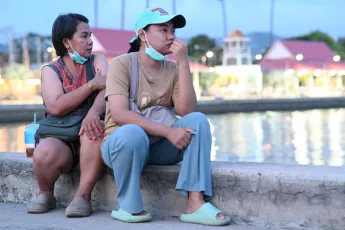 DSC_2298: Two women sit on a waterfront concrete ledge at dusk, masks lowered around their chins as they gaze toward the water, lost in thought.