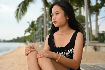 DSC_4534: A girl sits on a beachfront promenade, gazing toward the water with palms and a calm shoreline behind her.