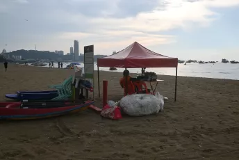 DSC_6170: A small red canopy set up on a sandy beach with kayaks and stacked chairs nearby, overlooking a calm sea and distant city skyline at dusk.