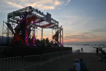 DSC_6229: A seaside concert stage glows with purple lights as the sun sets over the calm ocean, while a few people watch from the beach.