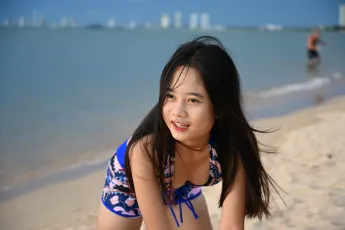 DSC_5065: A young girl in a colorful swimsuit smiling and leaning forward on a sandy beach with calm blue water and distant city buildings in the background.