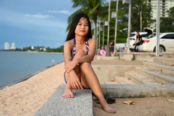 DSC_5124: Young woman in a colorful swimsuit sitting on a seawall, relaxing by the beach with palm trees and parked cars in the background.