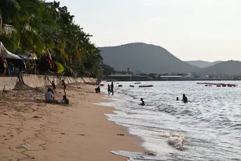 DSC_6921: Golden-hour beach scene with people relaxing on the sand and swimming near a palm-lined shore, hills rising in the distance.