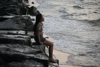 DSC_7254: A young woman sits on rocky shore, gazing peacefully at the waves rolling in.