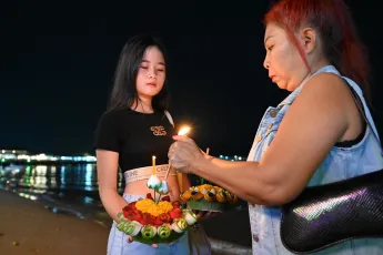 DSC_5938: Two women prepare and light floral krathongs by the water at night, ready to release them during a Loy Krathong celebration.
