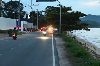 DSC_7780: Evening traffic along a coastal road as scooters and cars pass by the shoreline at dusk.