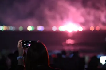DSC_6226: Someone in the crowd captures the dazzling pink glow of a distant fireworks display over the water on their phone.