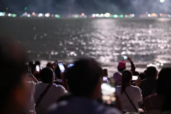 DSC_6205: A crowd of spectators by the water hold up phones and cameras, capturing a nighttime waterfront scene as lights shimmer across the surface.