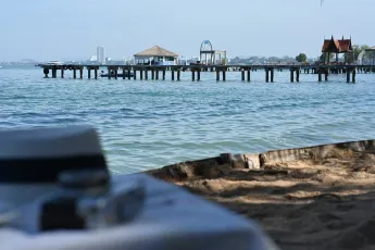 DSC_6310: A peaceful seaside pier stretching into calm blue waters, framed by sandy shore and a blurred camera in the foreground.