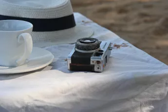 DSC_6308: Sipping sunlight and memories, a vintage camera and a straw hat rest beside a teacup on a sunlit tablecloth at the beach.