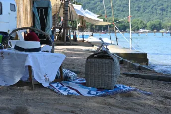 DSC_6311: A peaceful lakeside picnic setup with a wicker basket, blanket, and sun hat beside the water on a sandy shore.