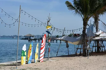 DSC_6305: Colorful surfboards rest on the sandy shore beneath string lights, with calm blue waters and a seaside pier in the background.