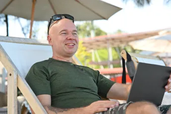 DSC_6336: A relaxed man lounges on a beach chair under an umbrella, smiling as he works on a laptop with sunglasses perched on his head.