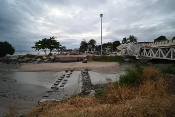 DSC_6691: A cloudy coastal scene with a sandy inlet and concrete steps leading to the water, a decorative bridge with a dragon-head detail on the right, and trees and buildings along the shoreline.
