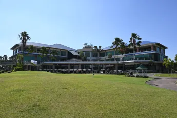 DSC_6921: A sunny day at a spacious beachfront resort and golf clubhouse, with palm trees lining the green lawn and a row of golf carts parked beneath the building's wide balconies.