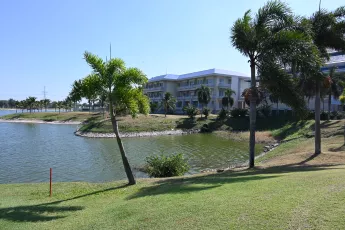 DSC_6918: Sunny waterfront view of a tropical resort complex with palm trees lining a calm lagoon and manicured green lawns.