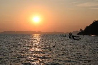 DSC_7638: Golden sun sinks over a tranquil sea, silhouetting boats and a distant shoreline as the water glitters with evening light.