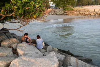 DSC_7753: A couple sits on sun-warmed rocks by the shoreline, talking as gentle waves lap the beach at golden hour.