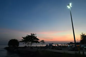 DSC_7775: A quiet seaside parking area at dusk, silhouetted trees and parked cars beneath a glowing streetlight as the sky fades from pink to deep blue.
