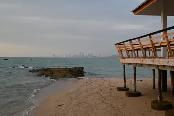 DSC_8985: A quiet beach at dusk with a weathered pier in the foreground and a distant city skyline hazy on the horizon.