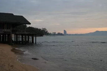 DSC_8986: A quiet shoreline at dusk with a stilted wooden structure in the foreground and a distant city skyline fading into the hazy horizon.