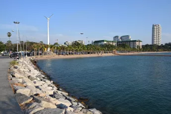 DSC_8242: A peaceful seaside promenade with rocky breakwater, calm blue water, palm-lined beach and modern buildings under a clear sky.