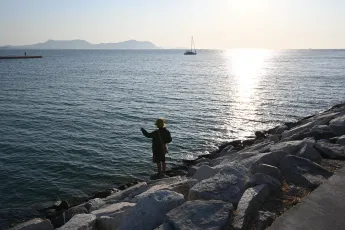 DSC_8237: A lone person stands on rocky shoreline, casting a line into calm, sunlit waters as a sailboat drifts on the horizon.