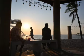 DSC_8260: Sunset silhouettes, people relax and stroll along the beach as lights and palm trees frame the golden horizon.