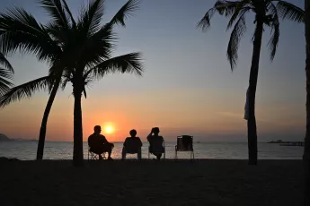 DSC_8266: Three friends silhouetted against a tropical sunset, relaxing under palm trees as the sun dips below the horizon.