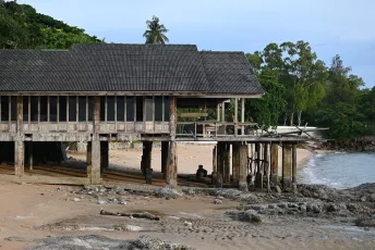 DFC_0460: Weathered wooden stilt house and dock built over a sandy, rocky shoreline with tropical trees in the background.