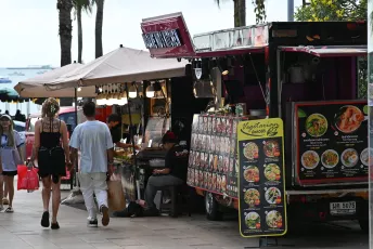 DFC_0849: A seaside food truck and street vendors line a palm‑shaded promenade as people stroll past, browsing menus and enjoying the waterfront atmosphere.