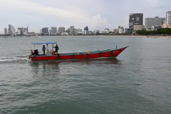 DFC_1176: A small red and green motorboat with three passengers cruises across calm coastal waters with a modern city skyline and sandy beach in the background.