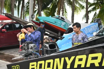 DFC_1297: Mechanics prepare powerboats on the beach, tuning engines and checking equipment under swaying palm trees.