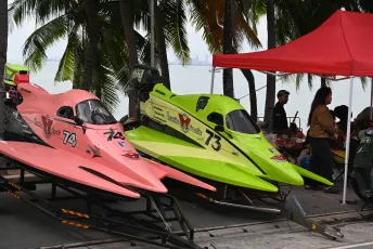 DFC_1298: Two brightly colored powerboats — one neon green numbered 73 and one pink numbered 74 — sit on trailers beneath palm trees and a red canopy at a waterfront event.