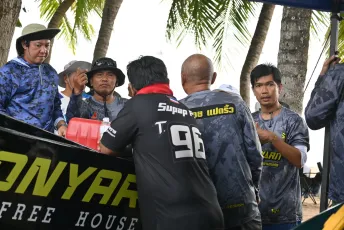 DFC_1411: A group of men in matching blue camo shirts and hats stand under palm trees, talking around a table with equipment and a banner as they prepare for an outdoor event.