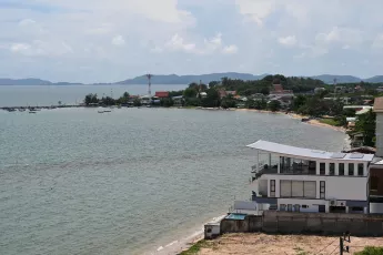 DZ6_1320: A peaceful coastal town with calm blue-green waters, a sandy shoreline, and modern waterfront homes overlooking distant islands on the horizon.