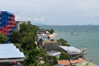 DZ6_1321: Coastal view of a seaside town with colorful buildings and green trees along the shoreline, small boats scattered on calm blue water, and a distant city skyline on the horizon.